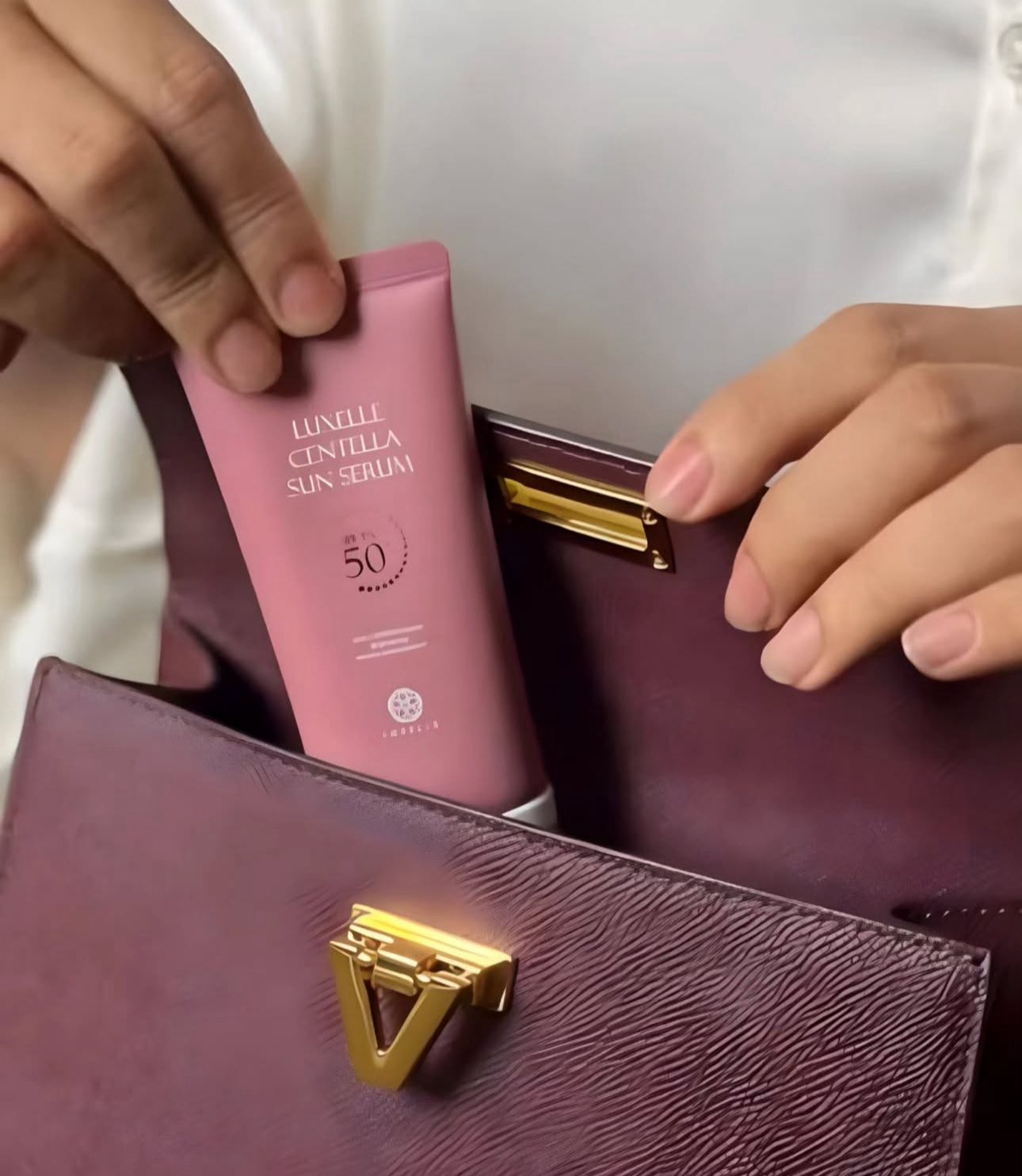 Pink sunscreen bottle with gold text held by a hand next to a purple handbag.
