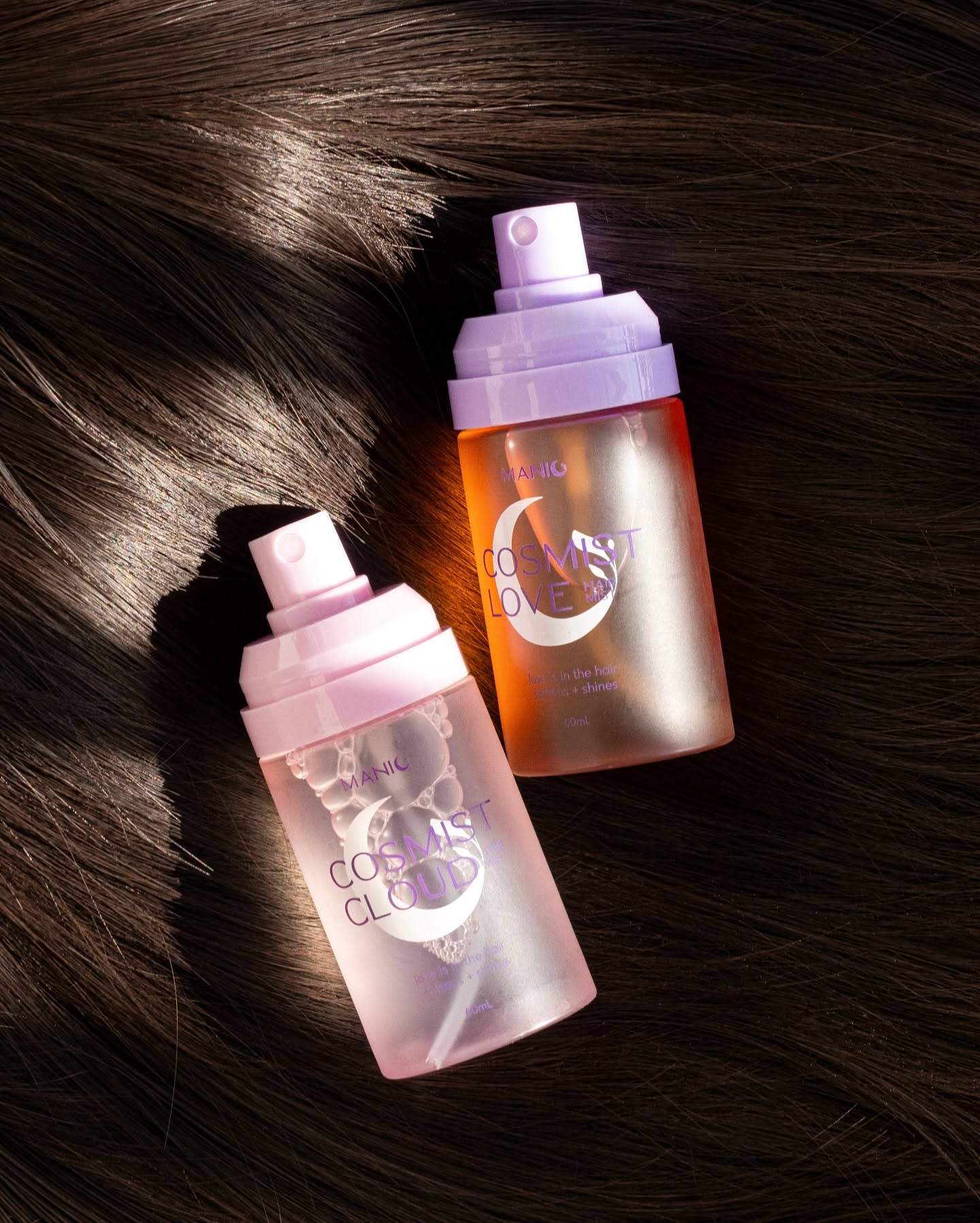 Two COSMOS CLOUD hair care spray bottles, one in translucent pink and one in translucent orange, displayed on a bed of dark brown hair.