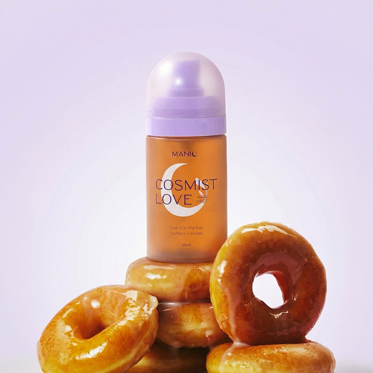 A bottle of MANIC Cosmist Love Dewy Setting Milk Makeup in a warm peachy-orange shade, styled on top of a stack of glazed donuts against a soft lavender background.