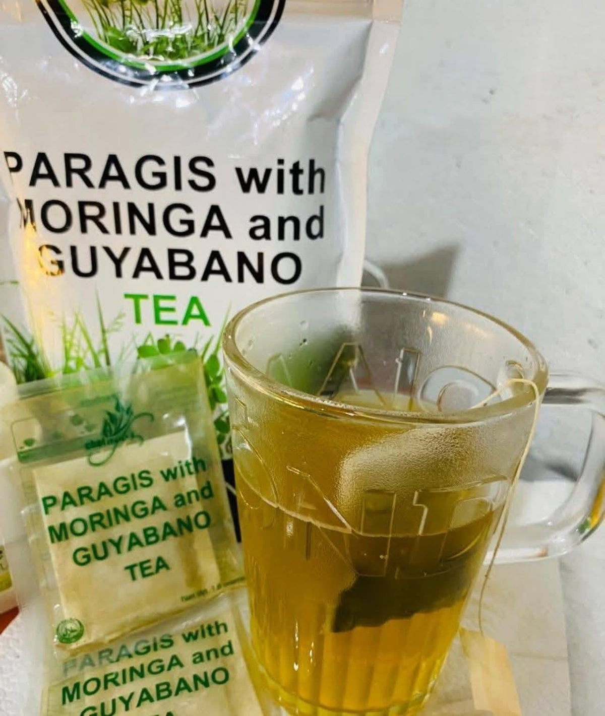 A glass mug filled with brewed tea in the foreground and a branded tea packet behind it. The visible text on both the packet and the mug reads 'PARAGIS with MORINGA and GUYABANO TEA'.