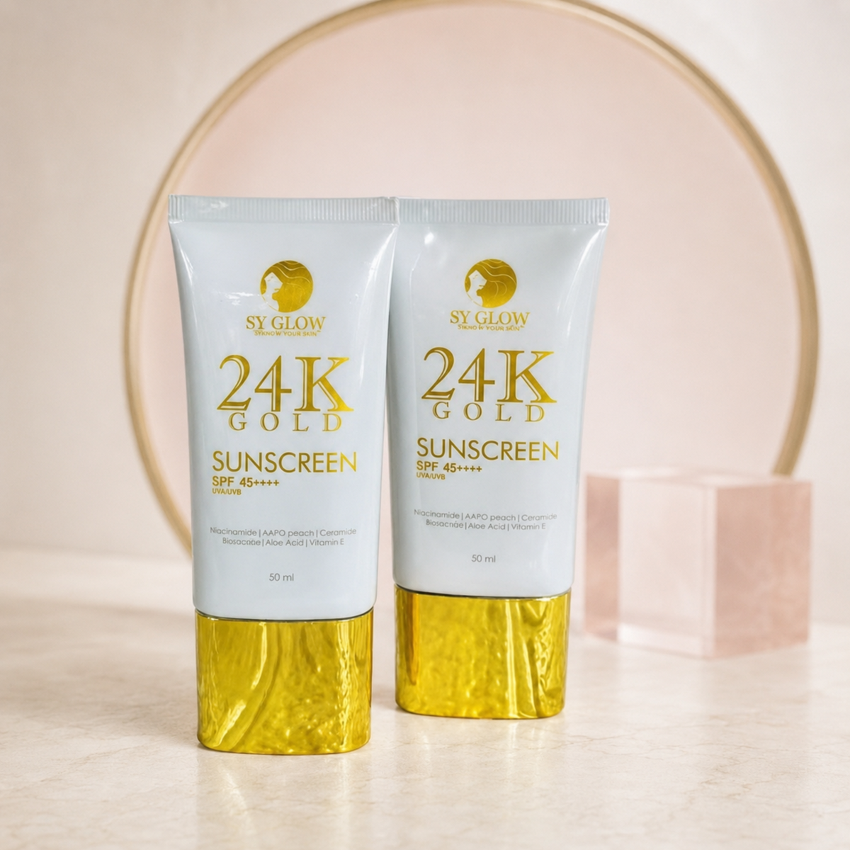 Two 50 ml tubes of SY GLOW 24K Gold Sunscreen SPF 45+ on a light marble surface with a gold circular backdrop and a pink block prop.