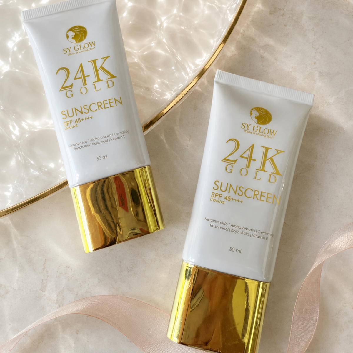 Two white tubes of SY GLOW 24K Gold Sunscreen SPF 48++++ with metallic gold caps, arranged on a light marble surface with a gold tray and ribbon.