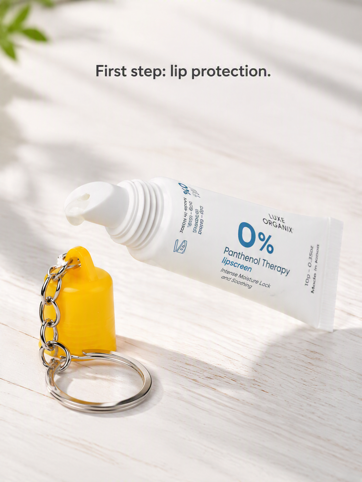 A white tube of Panthenol Therapy lipscreen by Suns Organix lies on a light wood surface next to a small yellow cap-shaped keychain with a metal ring. Text at the top reads, 'First step: lip protection.