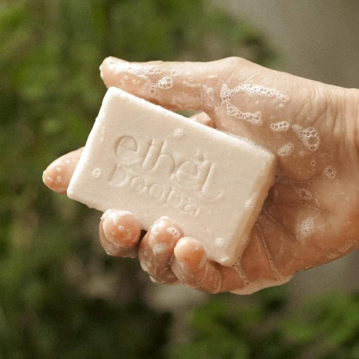A hand covered in soap lather holds a rectangular bar of Ethel Deoda soap, with a soft-focus green outdoor background.